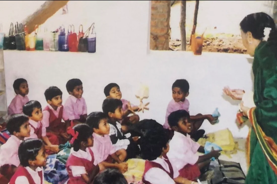 An old photograph of a teacher in a green saree with a class of young children in school uniforms sitting on the floor
