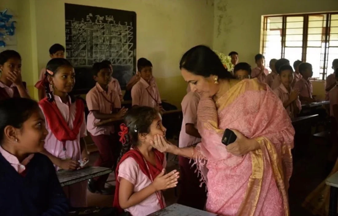 A woman in a pink saree lovingly holding the face of a young schoolgirl in a classroom