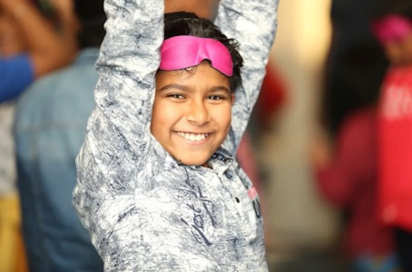 A smiling boy with his blindfold pushed up, arms stretched overhead