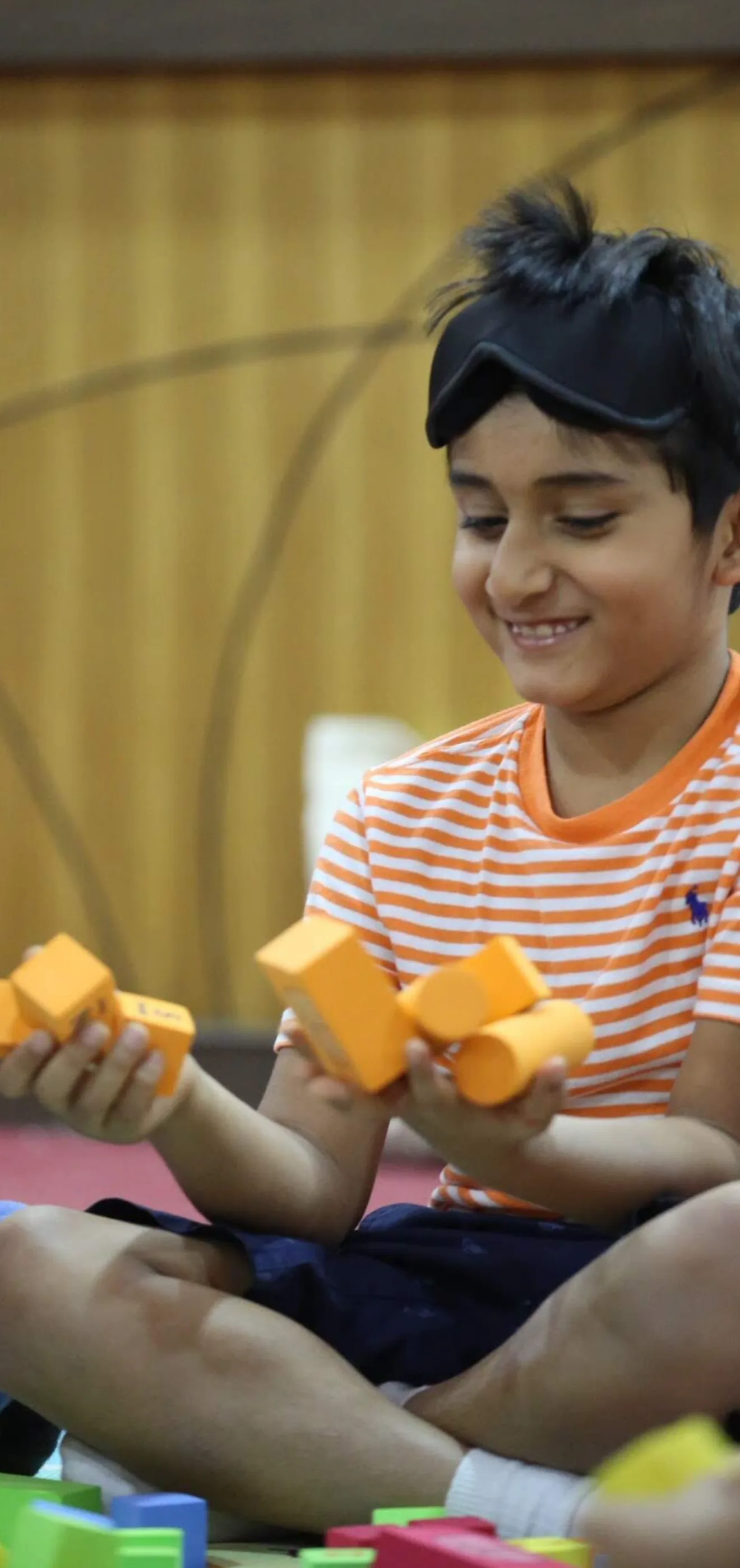 A smiling boy, eye mask pushed up, holding orange foam blocks