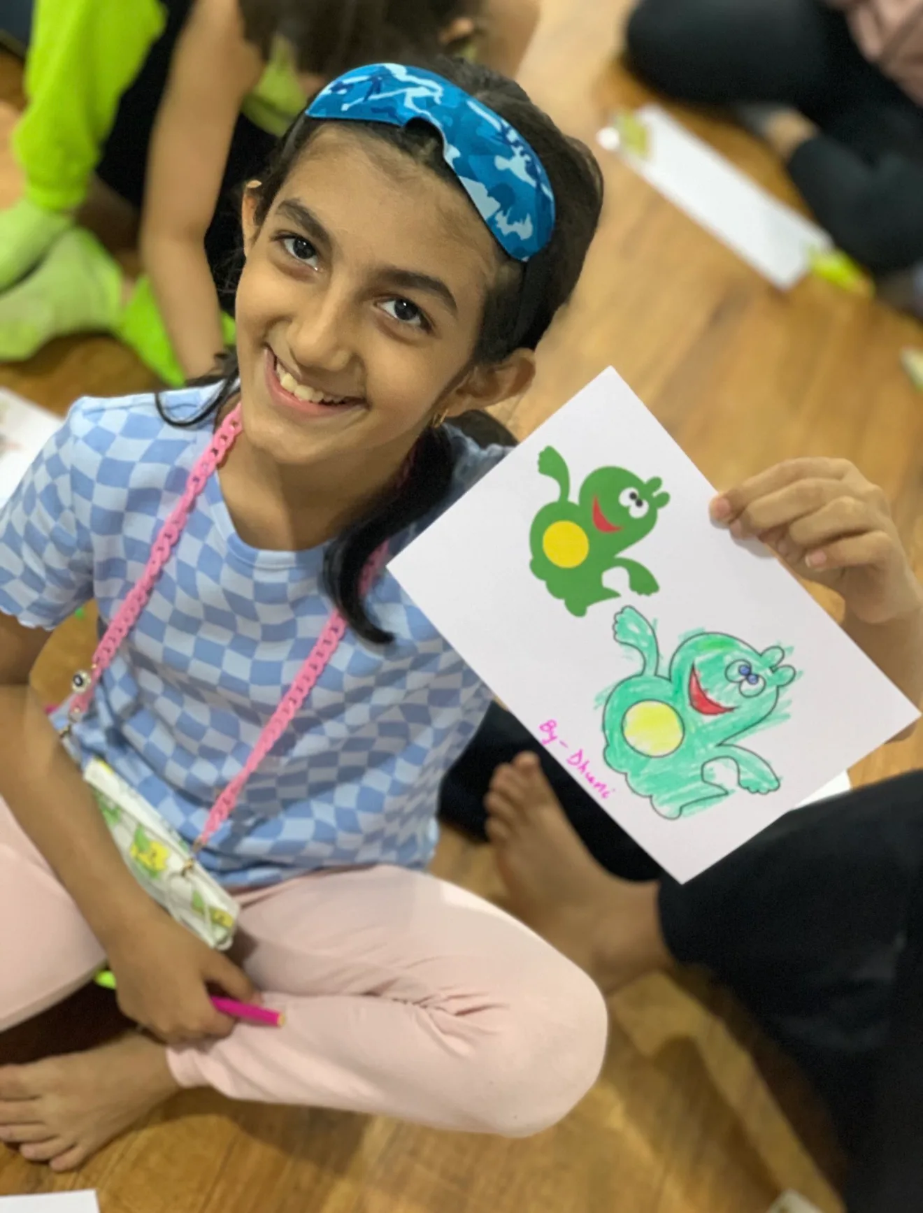 A smiling girl, blindfold pushed up, holding her crayon copy of a frog picture