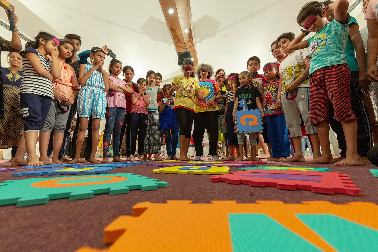 Blindfolded children searching for foam alphabet tiles on the floor while others watch
