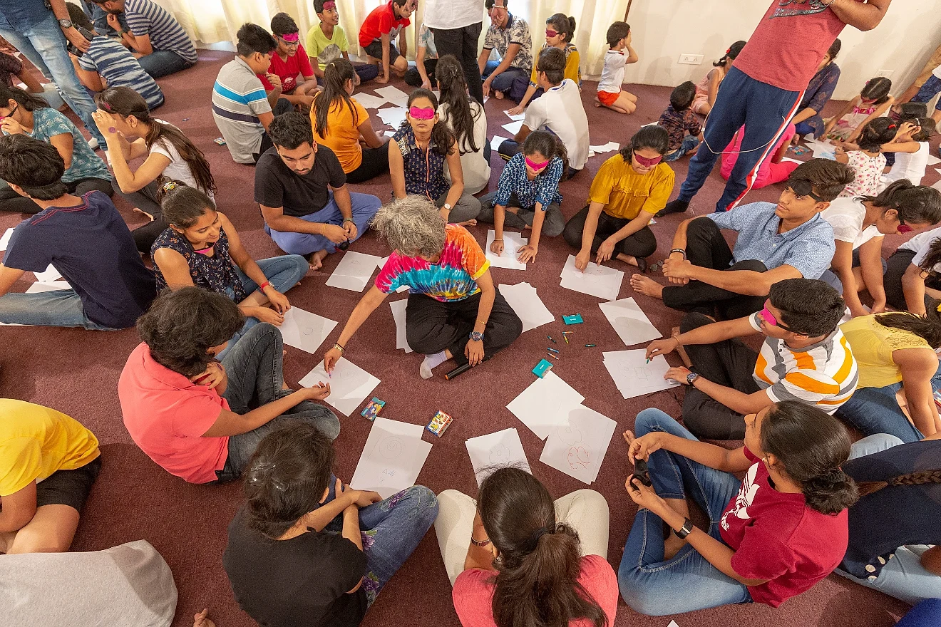 Shreya sitting on the floor at the centre of a circle of blindfolded children and teens, who are drawing on sheets of paper during the Intuition Process