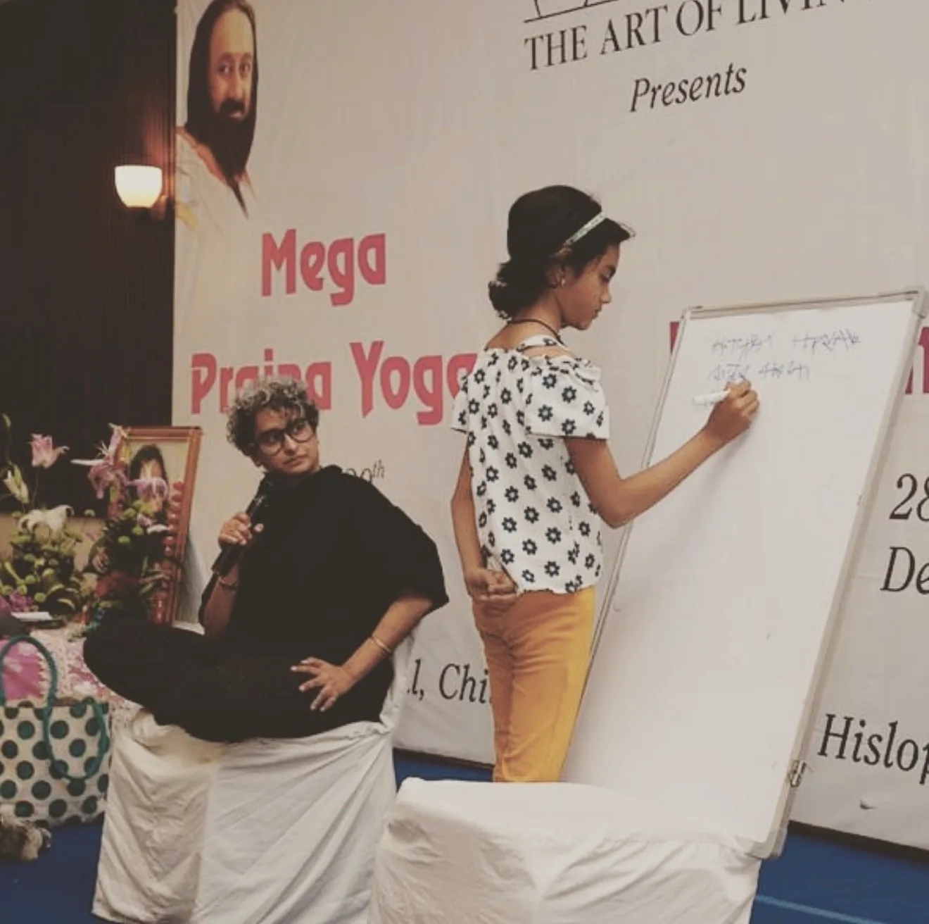A girl writing on a whiteboard on stage while Shreya looks on, at a Mega Prajna Yoga programme