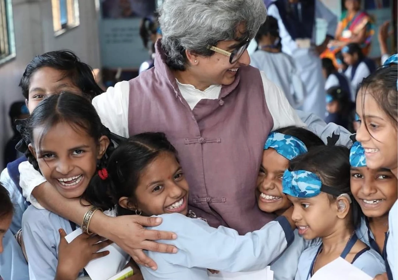 Shreya laughing as a group of smiling schoolgirls in uniform, some wearing blindfolds on their foreheads, crowd in for a hug