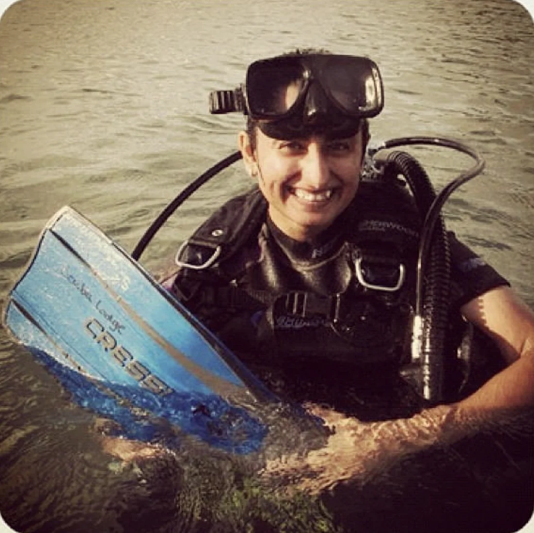Shreya smiling in the water in full scuba gear, holding a blue fin