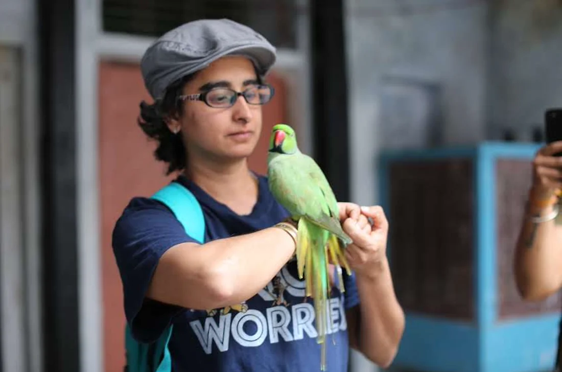 Shreya in a grey cap with a green parakeet perched on her hand