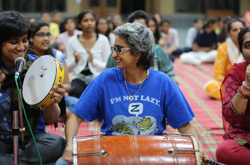 Shreya laughing as she plays a dholak at a satsang, with a friend on tambourine and a hall of people clapping