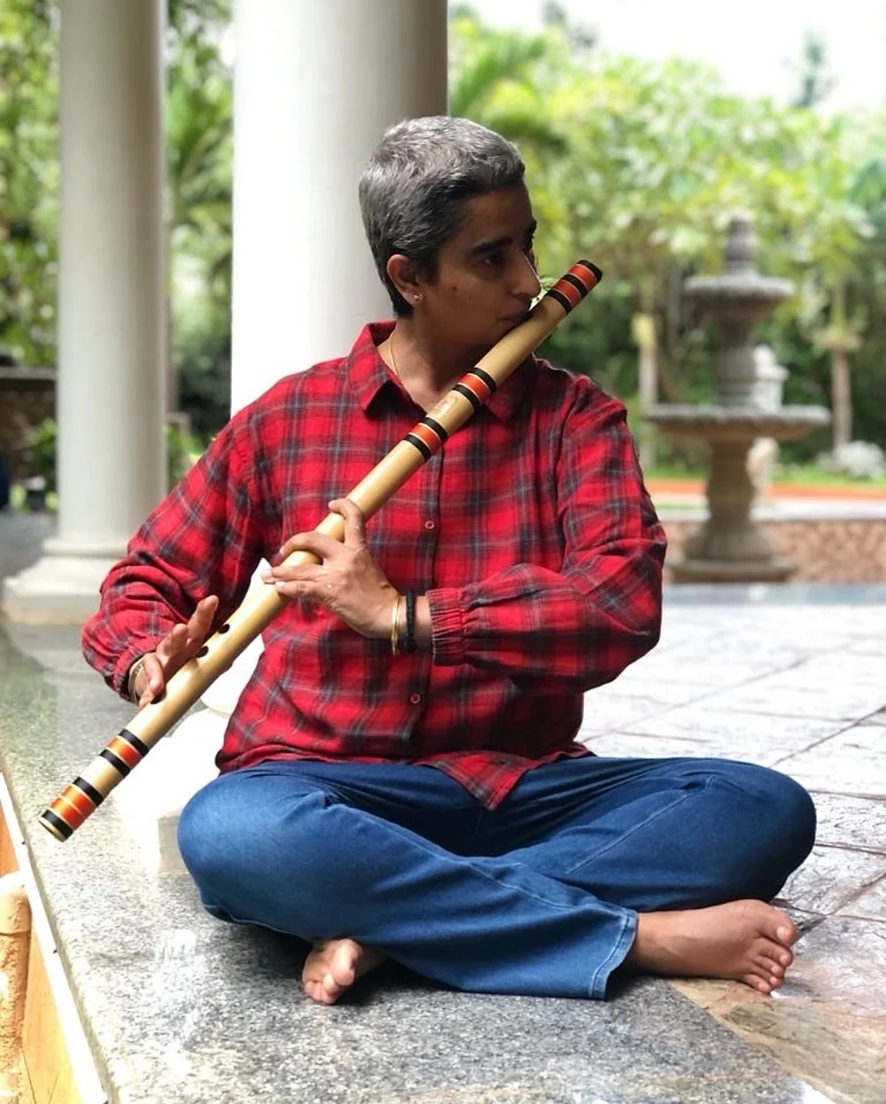 Shreya sitting cross-legged in a garden courtyard, playing a long bamboo flute