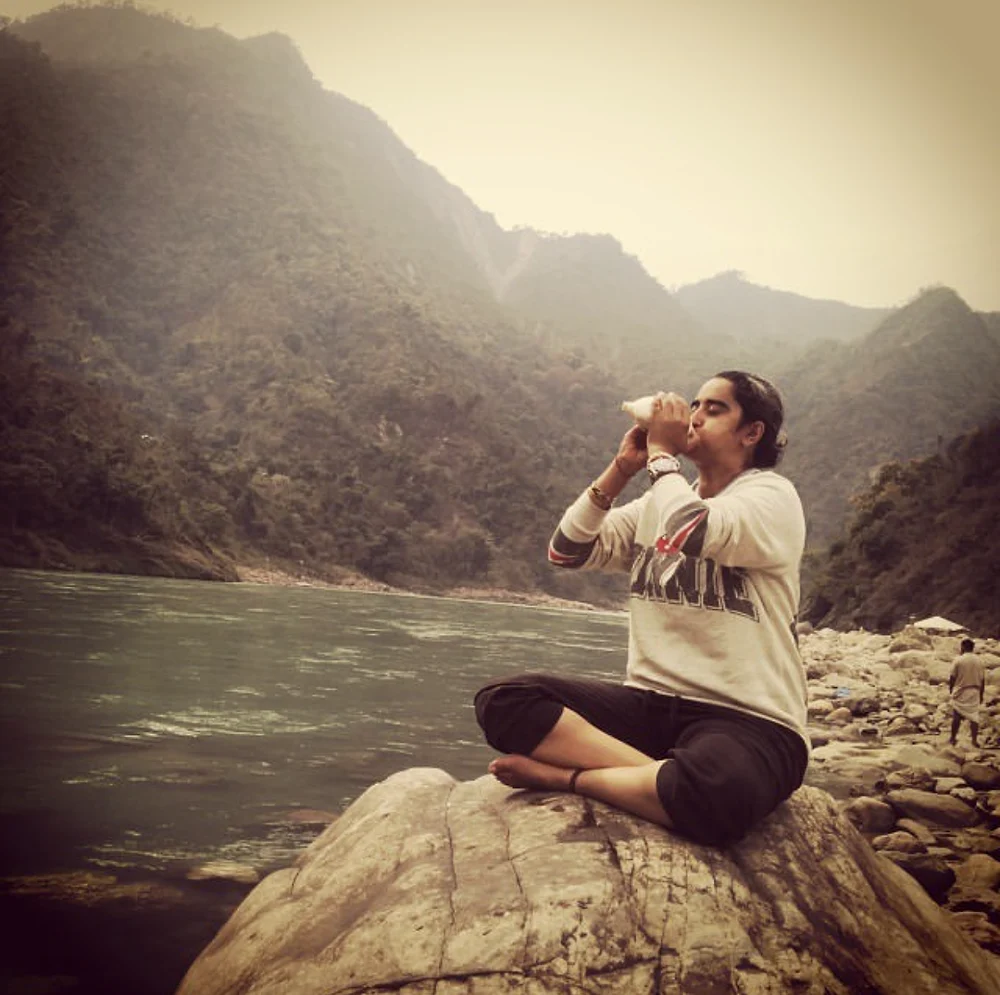 Shreya sitting cross-legged on a rock by the Ganga in Rishikesh, blowing a conch shell