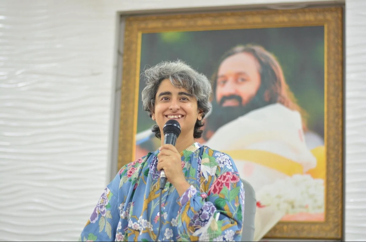 Shreya Chugh smiling as she speaks into a microphone, in front of a framed portrait of Gurudev Sri Sri Ravi Shankar