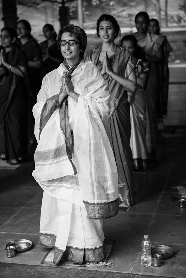 Shreya in a white veshti and angavastram standing with palms joined in namaskar, a row of women in saris behind her