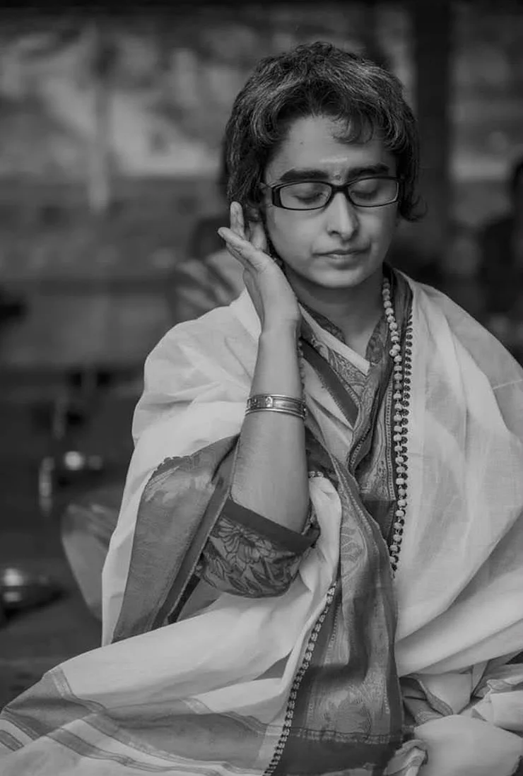 Shreya seated in white with eyes closed, one hand raised to her ear, a mala around her neck