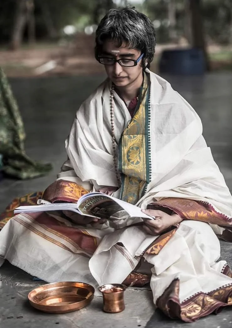 Shreya seated on the floor in white and gold, reading from a book, with a copper plate and tumbler beside her