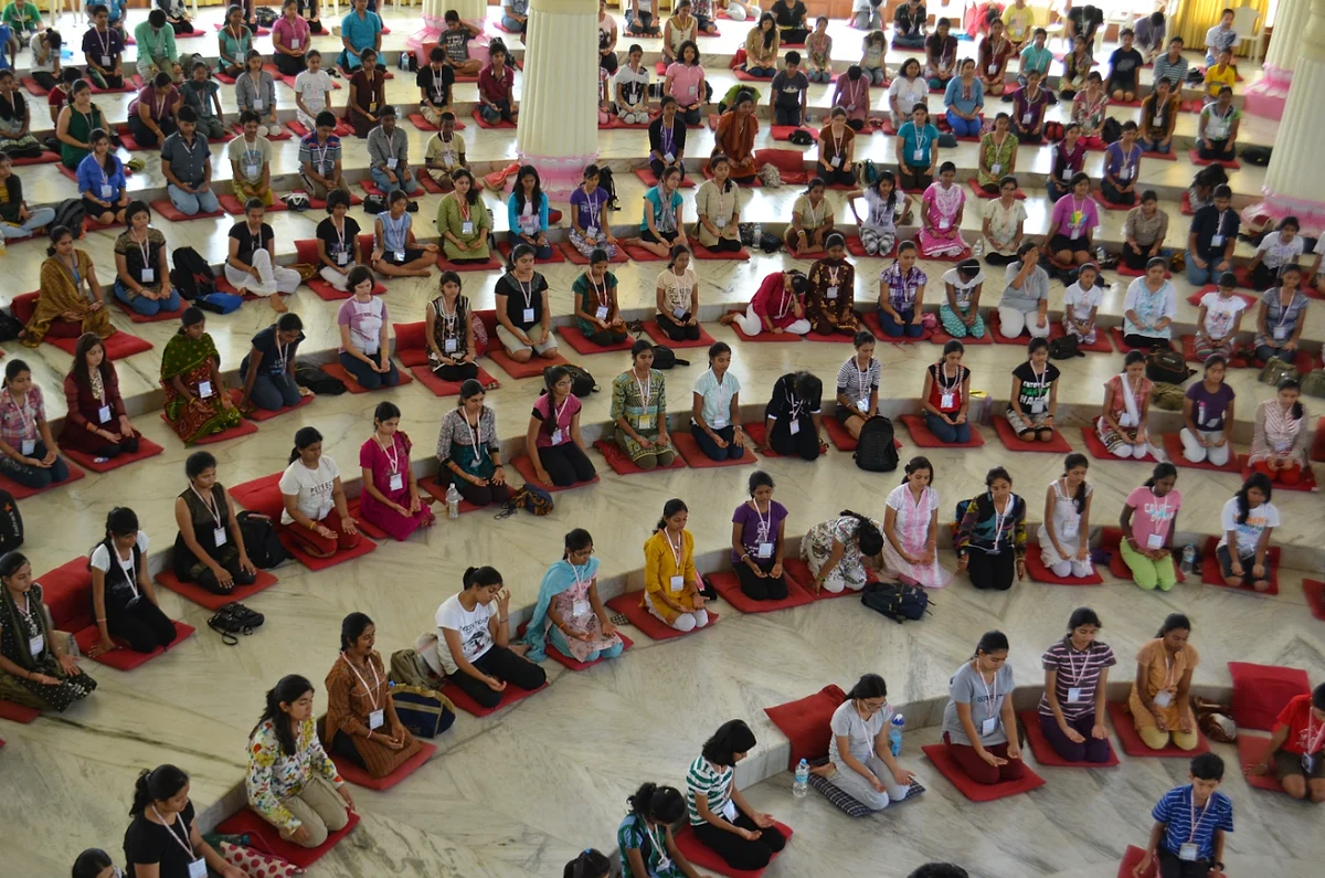Rows of young people seated on red cushions in a marble hall, eyes closed in meditation
