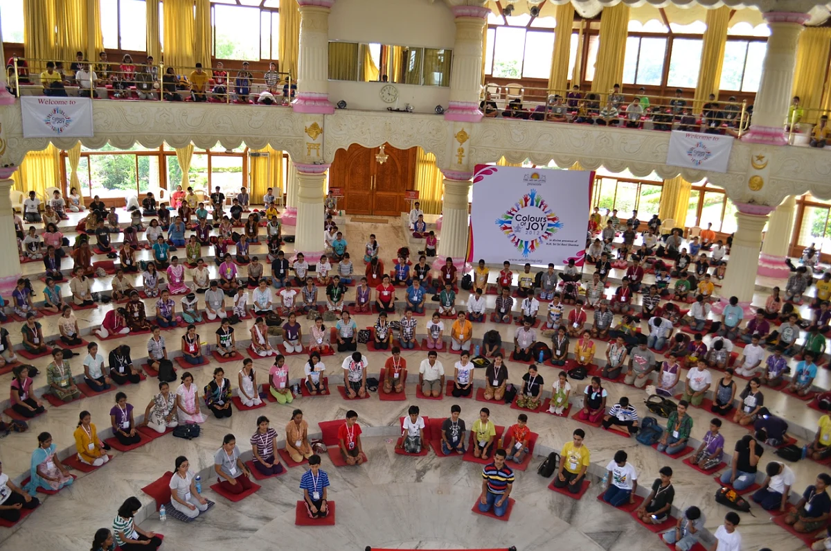 A vast round meditation hall filled with hundreds of teens seated in concentric circles on red cushions, under a Colours of Joy banner