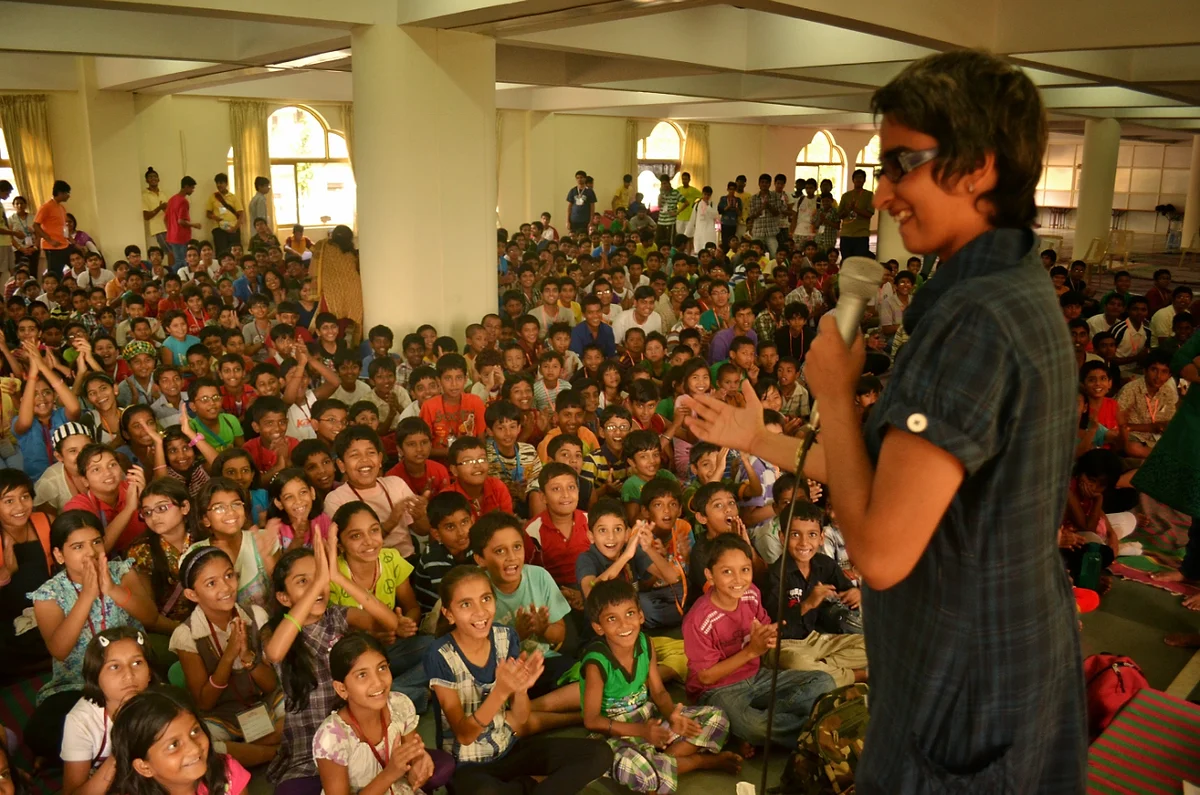 Shreya holding a microphone, laughing with a packed hall of cheering children